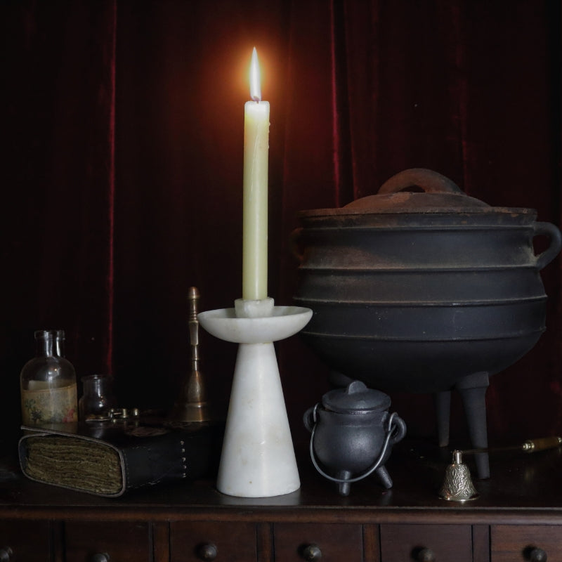 lit beeswax dinner candle in a white marble candle holder. In the background there is a small and a large cauldron, a black book of shadows , dark brass altar bell, an ornate brass candle snuffer and apothecary jars with floral labels. All items are sitting on a wooden apothecary cabinet .