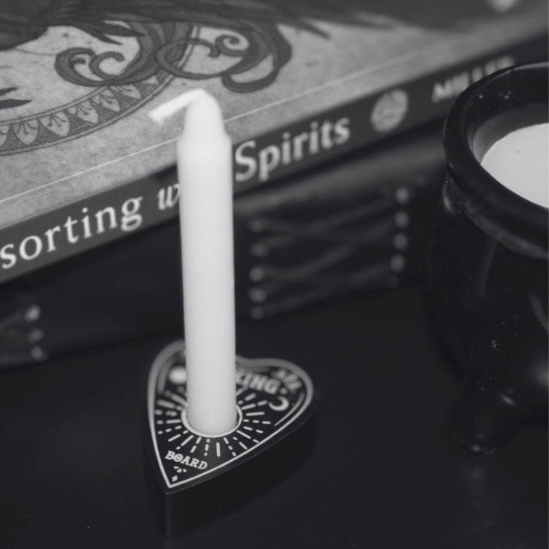 Black spell candle holder shaped as a Ouija board planchette, holding  a white spell candle. In front of a book "consorting with spirits" , leather journal and black ceramic cauldron candle