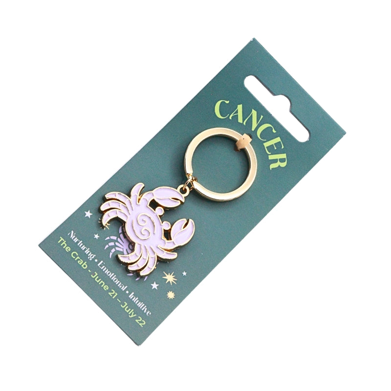A Cancer zodiac sign keychain featuring a pink and white enamel crab with gold-tone metal trim, attached to a gold-tone split ring and displayed on a dark green card.