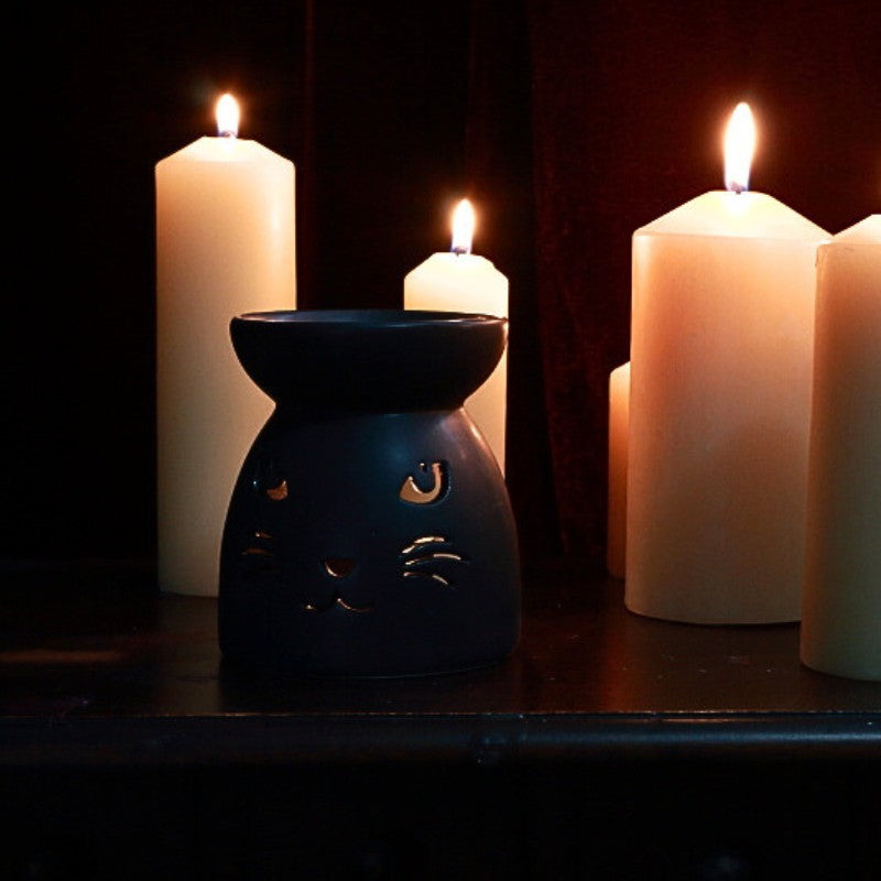 black oil burner with cut out of a cat's face on front. Lit beeswax pillar candles in the background.