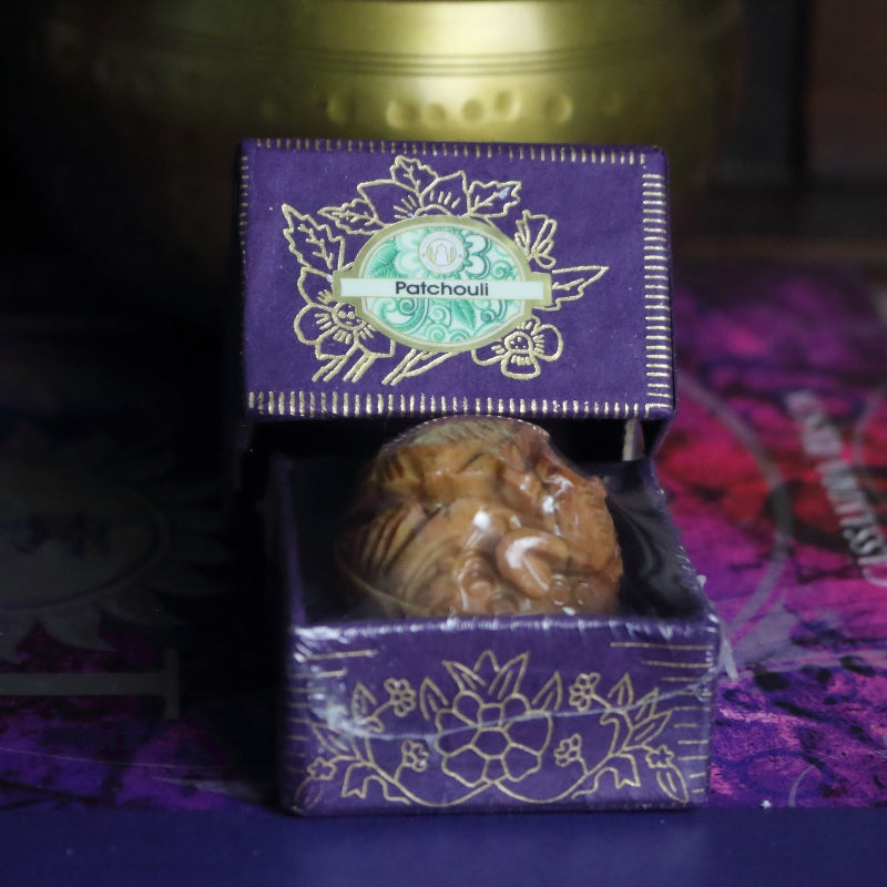 purple and gold box containing a hand carved soapstone jar, sitting on a purple and pink book of spells in front of a brass singing bowl with wooden striker.