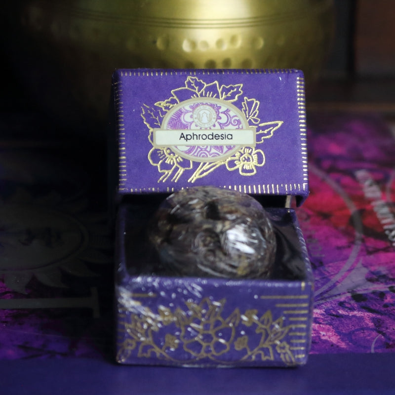 purple and gold box containing a hand carved soapstone jar, sitting on a purple and pink book of spells in front of a brass singing bowl with wooden striker.