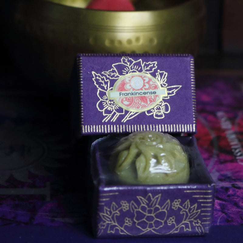 purple and gold box containing a hand carved soapstone jar, sitting on a purple and pink book of spells in front of a brass singing bowl with wooden striker.