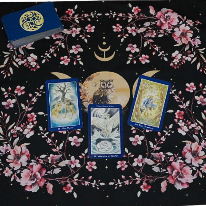 black tarot cloth with gold triple moon and owl design with floral borders. 3 card spread is laid out on top and a tarot deck in the corner