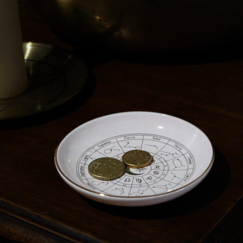 White dish with astrological design and coins on a dark surface