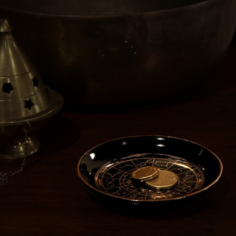 Decorative black dish with gold design and coin, next to a metallic cone-shaped object on a dark background.