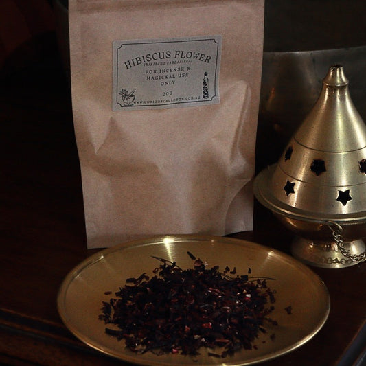 Hibiscus flower package with a brass incense burner and dish of dried flowers on a dark surface.