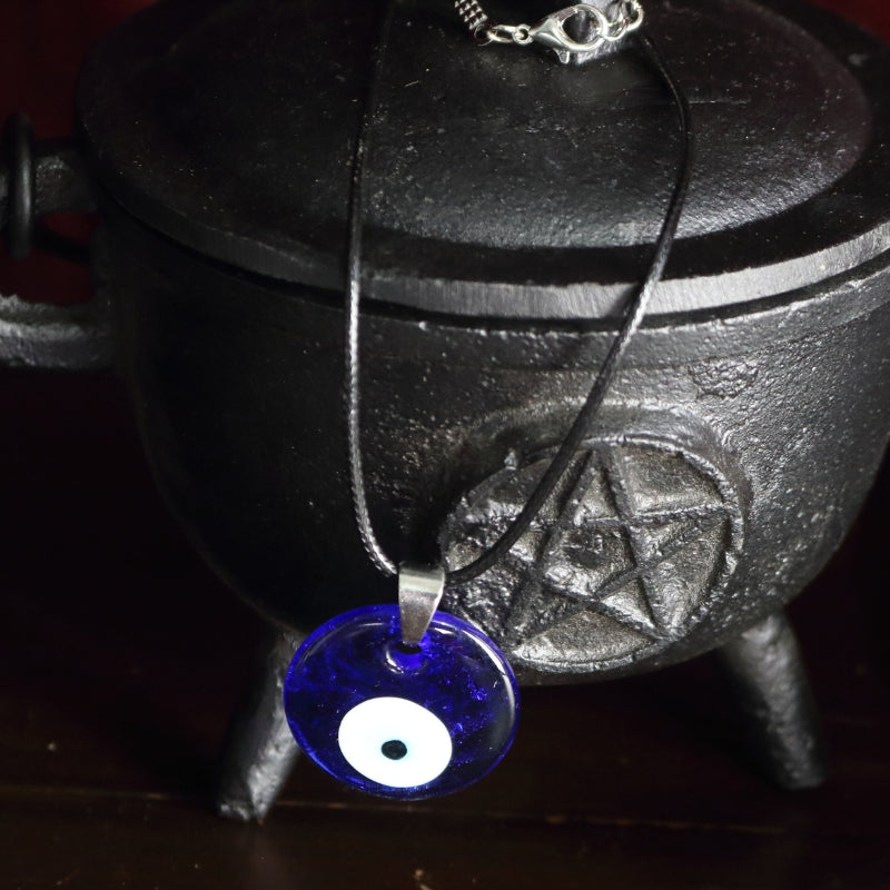 blue, white, light blue and black concentric circles formed into an eye shaped bead, hung on a black cord with a silver clasp. The necklace hangs from a black cast iron cauldron with a pentacle on the front of it.