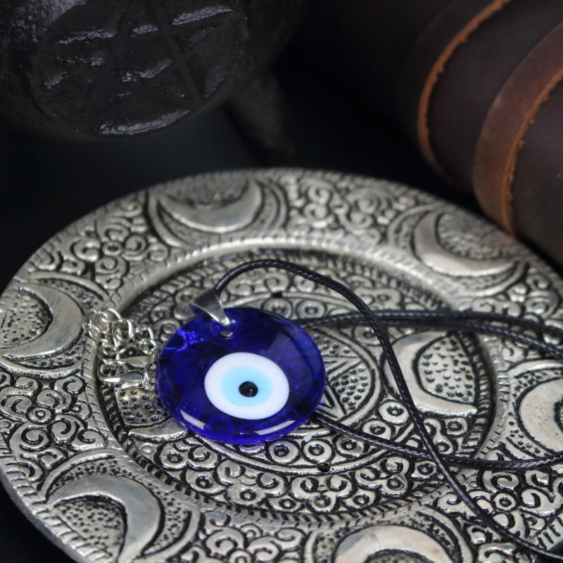 blue, white, light blue and black concentric circles formed into an eye shaped bead, hung on a black cord with a silver clasp. The necklace sits on an ornate silver plate with moons embossed on it, in front of a black cast iron cauldron with a pentacle on the front of it.