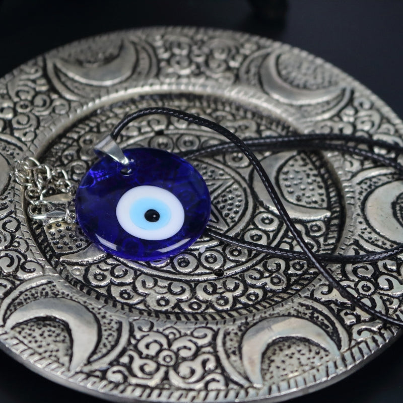 blue, white, light blue and black concentric circles formed into an eye shaped bead, hung on a black cord with a silver clasp. The necklace sits on an ornate silver plate with moons embossed on it.