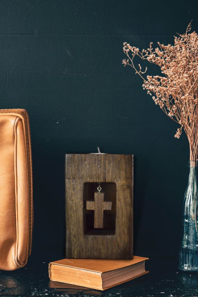 cross wooden tealight candle holder  solid hardwood with white tea light in top on a book, next to a leather bag and vase containing dried flowers, in front of a dark blue wall