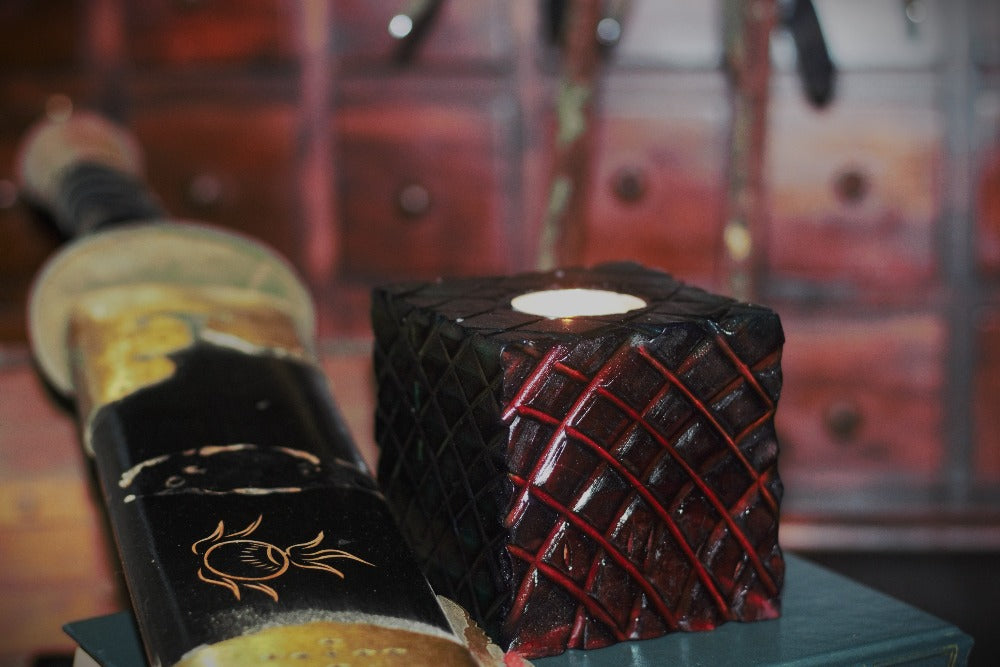 Red wooden square candle holder with diagonal lines etched to resemble dragon scales. Containing a white tea light candle, on a book next to a sword, in front of an apothecary chest