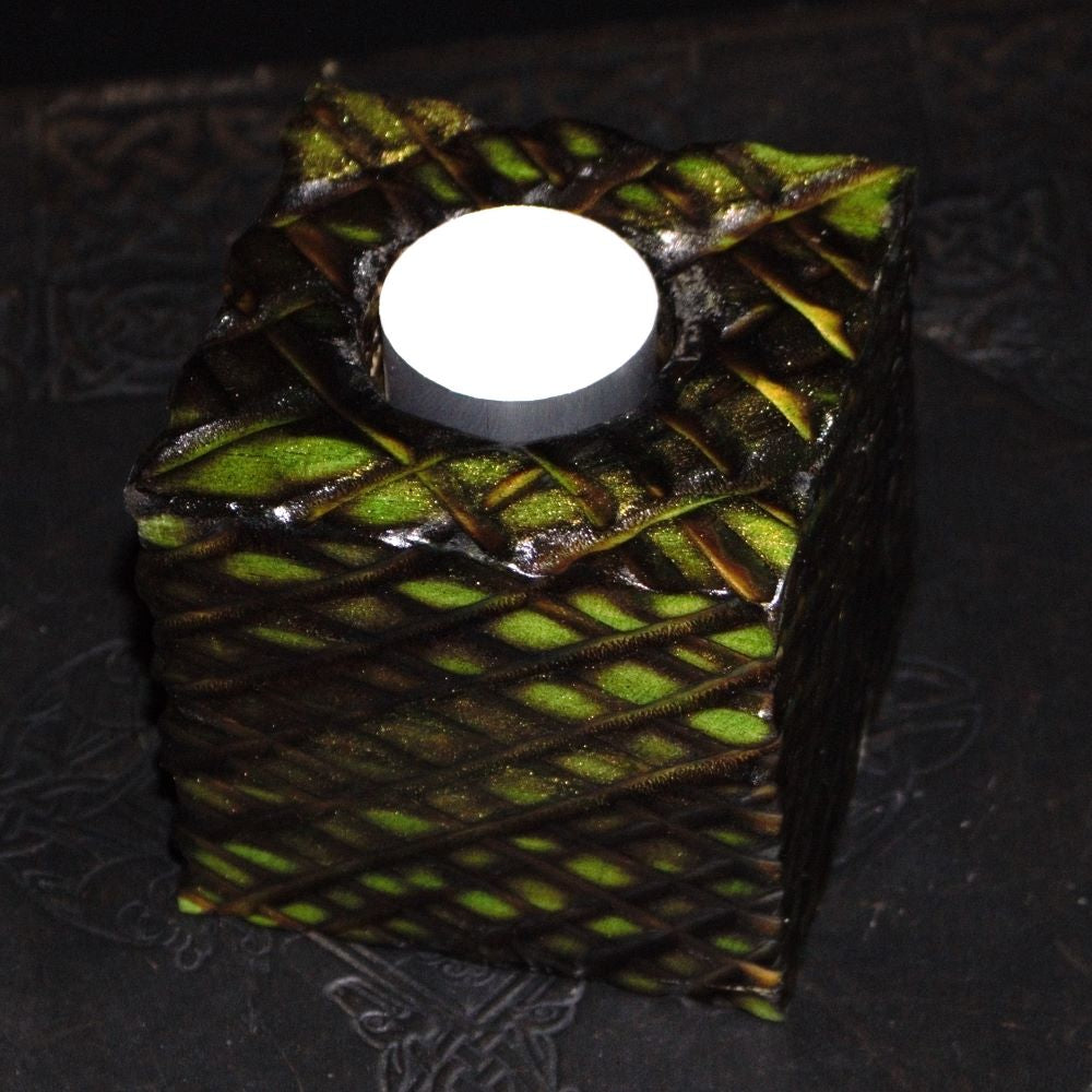 green and gold wooden square candle holder with diagonal lines etched to resemble dragon scales. Containing a white tea light candle, on a black embossed background