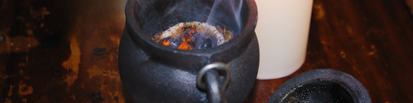 black cast iron cauldron with a lit charcoal disc inside,  sitting on a wooden table in front of a white candle