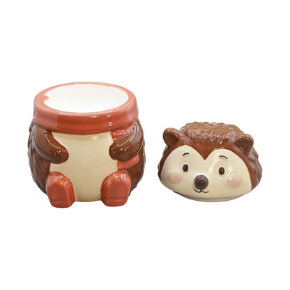 A small ceramic hedgehog-themed oil burner with a matching round lid placed beside it on a plain white background. The body is cream-colored with brown sides and feet, and the lid shows a brown hedgehog face with pink cheeks and a dark nose.