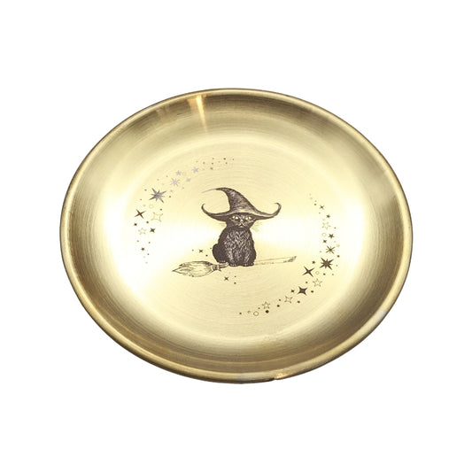 A round, brushed gold metal tray or shallow dish featuring a central illustration of a black owl wearing a witch's hat on a broomstick, surrounded by small stars.