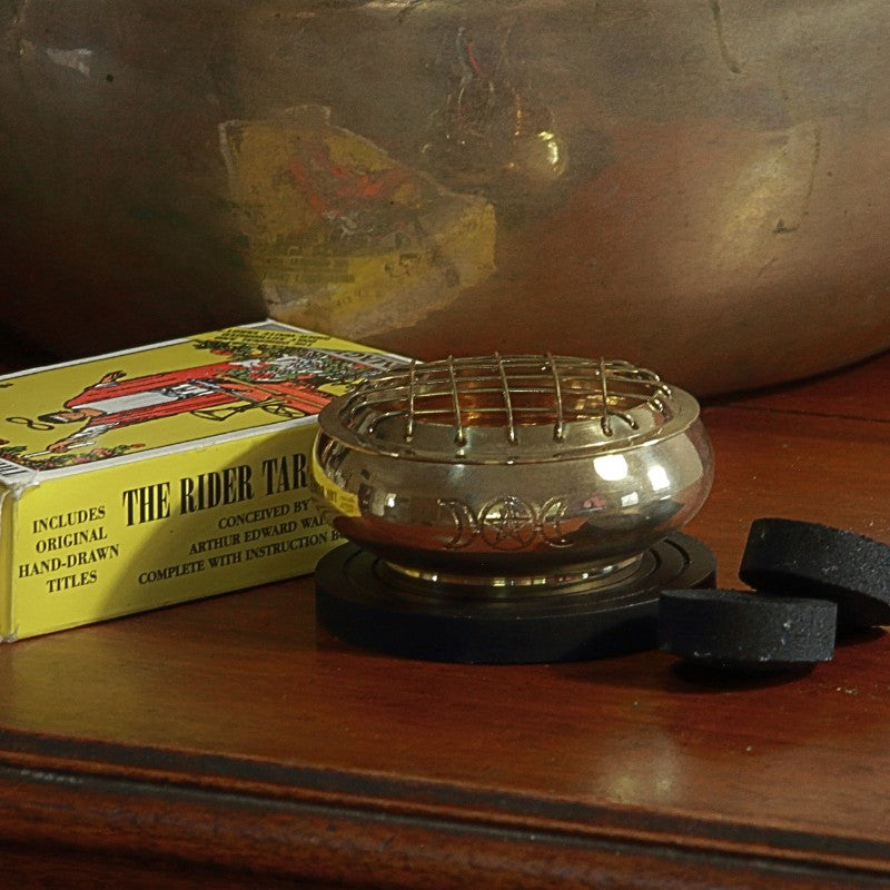 Brass charcoal incense burner engraved with the triple moon pentacle design, sitting on a wooden base plate next to charcoal discs and a rider waite smith tarot deck