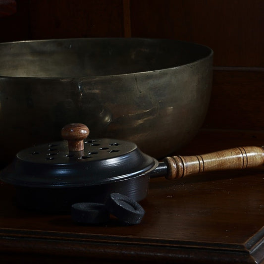 iron charcoal incense burner with lid and wooden handle next to a brass singing bowl and charcoal discs