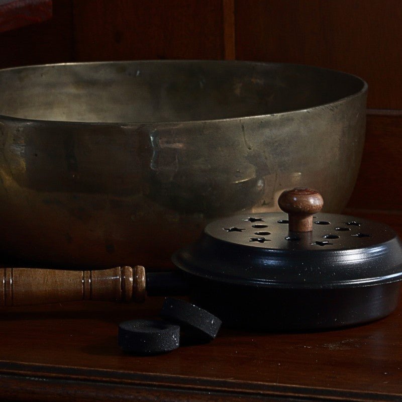 iron charcoal incense burner with lid and wooden handle next to a brass singing bowl and charcoal discs