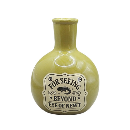 A small round ceramic vase with a short neck, in a glossy olive green glaze, featuring a vintage-style cream label with the text 'FOR SEEING BEYOND EYE OF NEWT' and a black newt illustration.