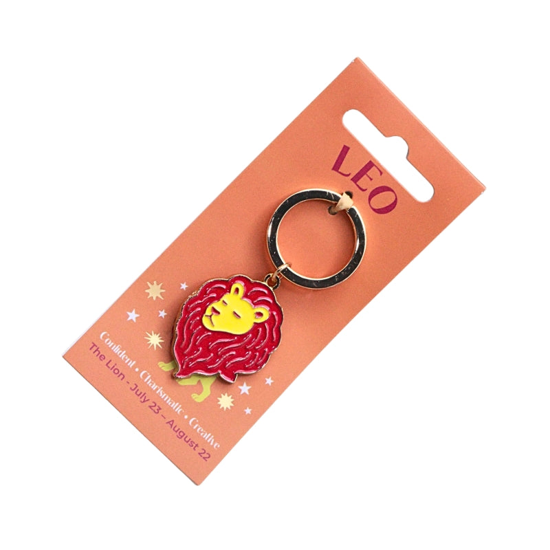 A Leo zodiac sign keychain featuring an enamel lion head charm with a red mane and yellow face, attached to a black split ring and displayed on a peach-colored cardboard backing card.
