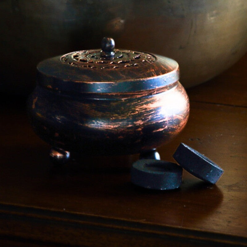 black and bronze coloured, round metal charcoal burner in front of a brass singing bowl next to charcoal tablets