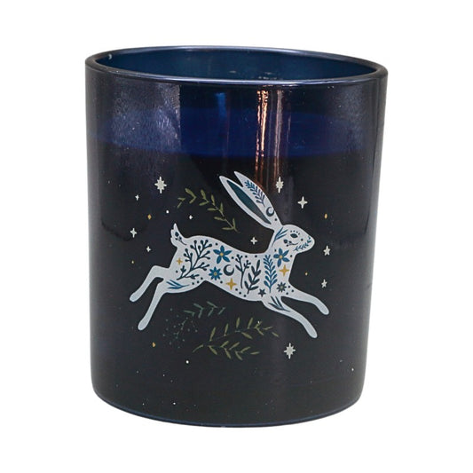 A dark navy blue glass candle jar featuring a white line-art illustration of a hopping rabbit decorated with blue and green floral patterns, surrounded by small stars and leaves.