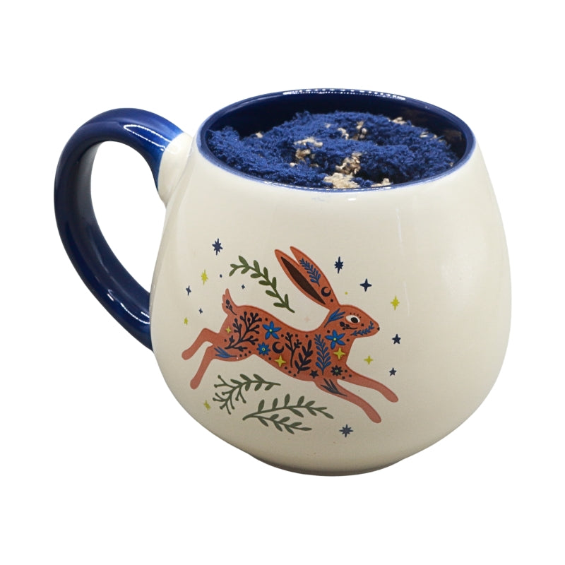 A ceramic mug with a cream-colored exterior, a dark blue handle and interior, and a hand-painted hopping rabbit design in orange, green, and blue on the front.