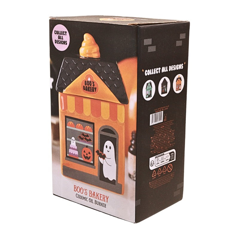 A boxed ceramic oil burner shaped like a Halloween-themed bakery storefront, shown on a plain white background. The packaging features orange and black with a ghost and pumpkin illustration on the front.
