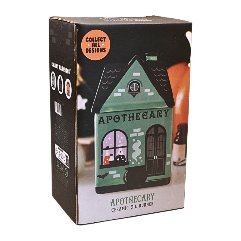 A boxed ceramic oil burner shaped like a haunted house labeled 'APOTHECARY.' The packaging is mostly black with a printed photo of the green house and an orange circular badge reading 'Collect All Designs.