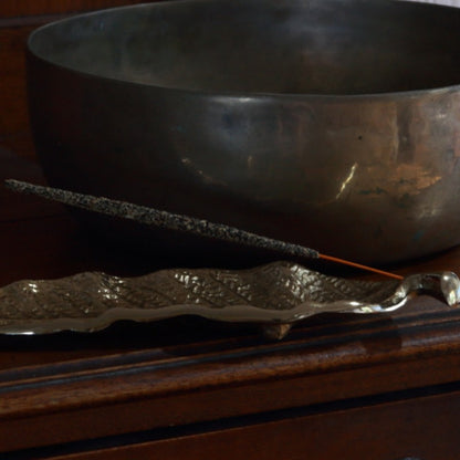 Handmade incense stick in a gold leaf incense stick holder, in front of a brass singing bowl on a wooden table