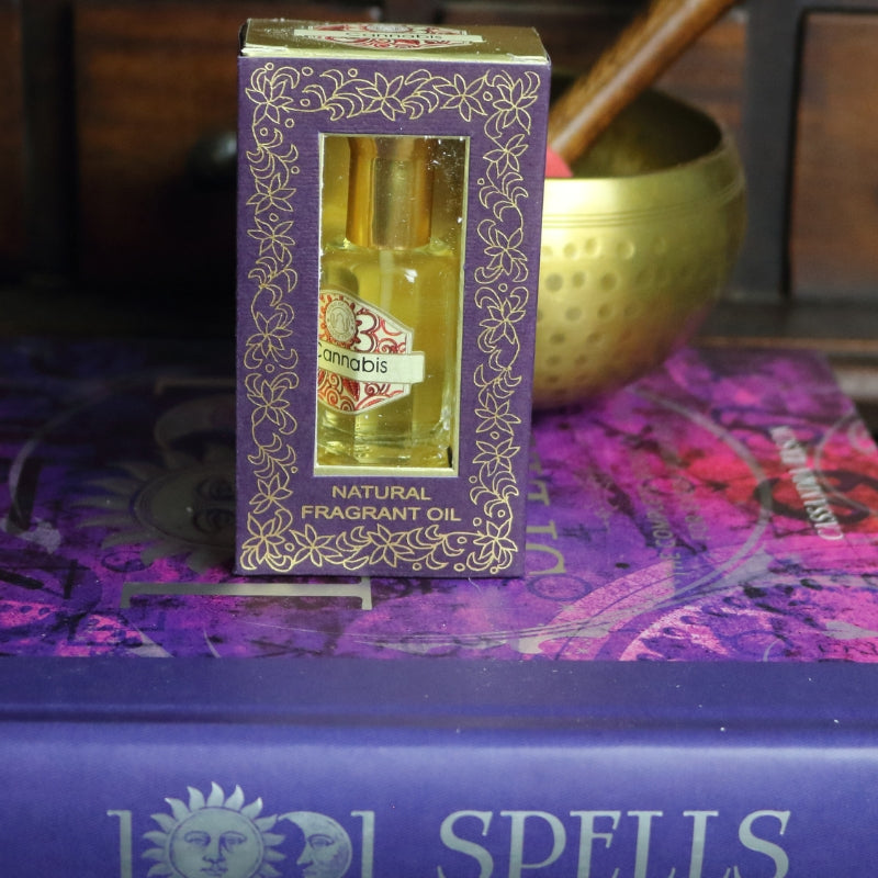 purple and gold box containing a miniature perfume bottle, sitting on a purple and pink book of spells in front of a brass singing bowl with wooden striker. in the background there is an apothecary cabinet