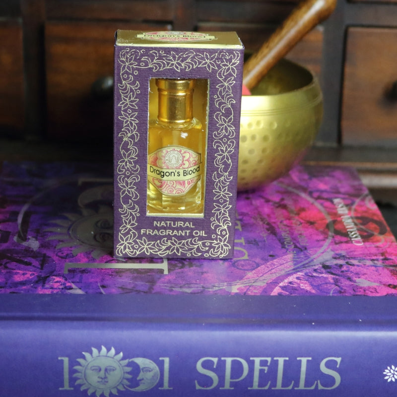 purple and gold box containing a miniature perfume bottle, sitting on a purple and pink book of spells in front of a brass singing bowl with wooden striker. in the background there is an apothecary cabinet