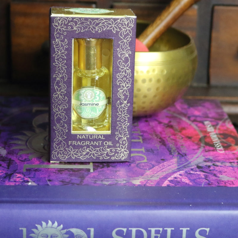 purple and gold box containing a miniature perfume bottle, sitting on a purple and pink book of spells in front of a brass singing bowl with wooden striker. in the background there is an apothecary cabinet