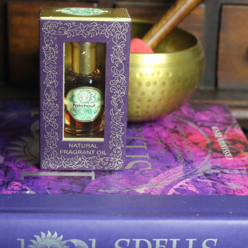 purple and gold box containing a miniature perfume bottle, sitting on a purple and pink book of spells in front of a brass singing bowl with wooden striker. in the background there is an apothecary cabinet