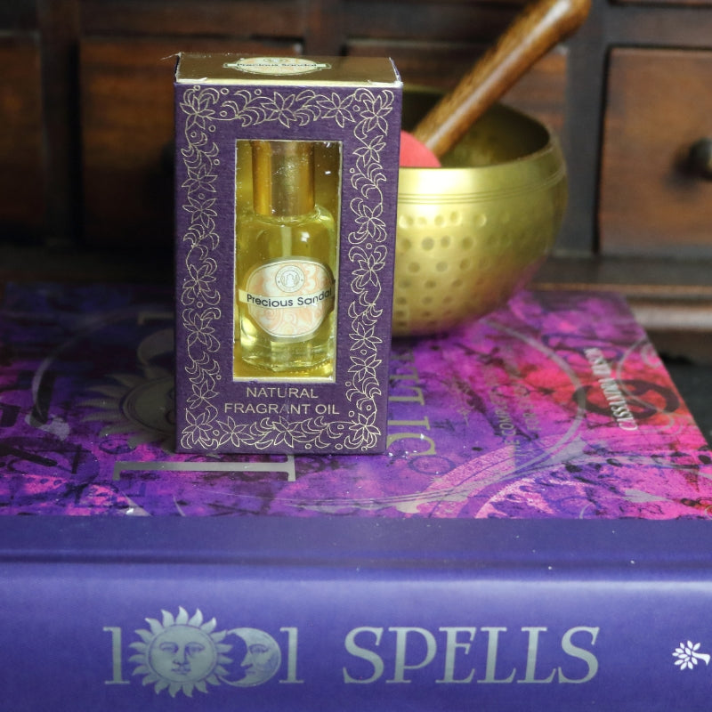 purple and gold box containing a miniature perfume bottle, sitting on a purple and pink book of spells in front of a brass singing bowl with wooden striker. in the background there is an apothecary cabinet