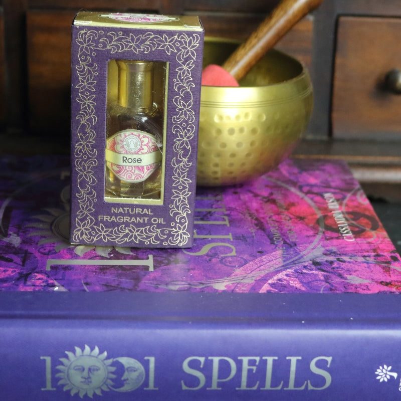 purple and gold box containing a miniature perfume bottle, sitting on a purple and pink book of spells in front of a brass singing bowl with wooden striker. in the background there is an apothecary cabinet