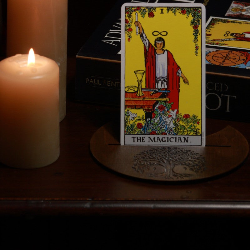 handmade tarot card holder with tree of life design, holding a rider waite magician tarot card, in front of a tarot book with candles in the background