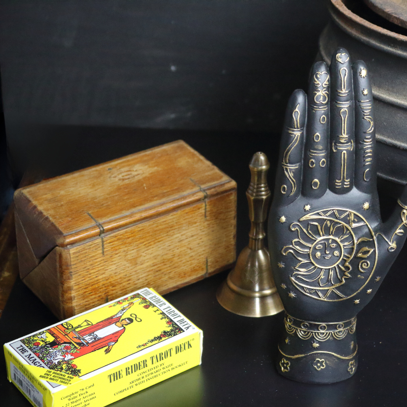 rider waite tarot deck sitting in front of a wooden tarot box next to an old cauldron, brass altar bell and black carved hand with gold symbols on its palm.