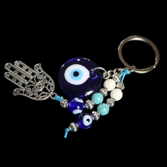 silver key ring attached to 2 beaded cords of white, light blue and evil eye beads (dark blue beads with a white centre and light blue and black circles inside. One of the beaded strings is attached to a Silver hamsa hand . A silver chain attached to a  blue bead with a white and black centre, on a black background