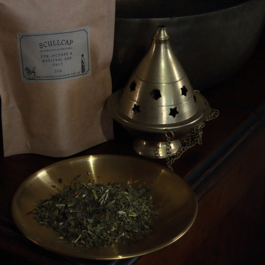 Brass bowl with dried herbs, brass cone incense burner, and Scullcap packaging on a wooden surface.