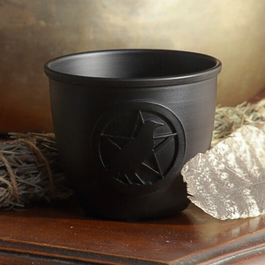 Iron smudge bowl with a raven pentacle design on front on a wooden table with a rosemary smudge stick and feather
