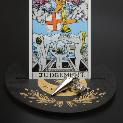 silver conical pendulum sitting on a moon shaped piece of wood painted black with a crescent moon on it, with a groove cut down the middle of it, holding a judgement rider waite tarot card.