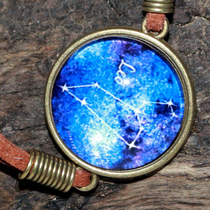 leather bracelet with a blue glass cabochon bead in the centre with the line drawing of the constellation of leo and the word leo written in white. The bracelet hangs on a piece of driftwood. 