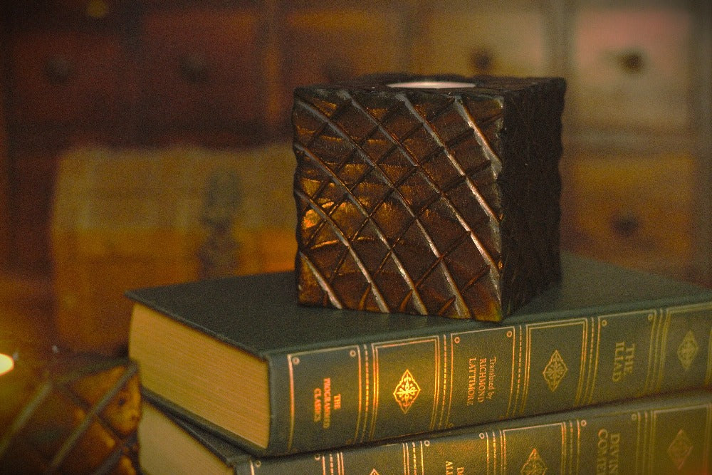 Red wooden square candle holder with diagonal lines etched to resemble dragon scales. Containing a white tea light candle, on a stack of books , in front of an apothecary chest