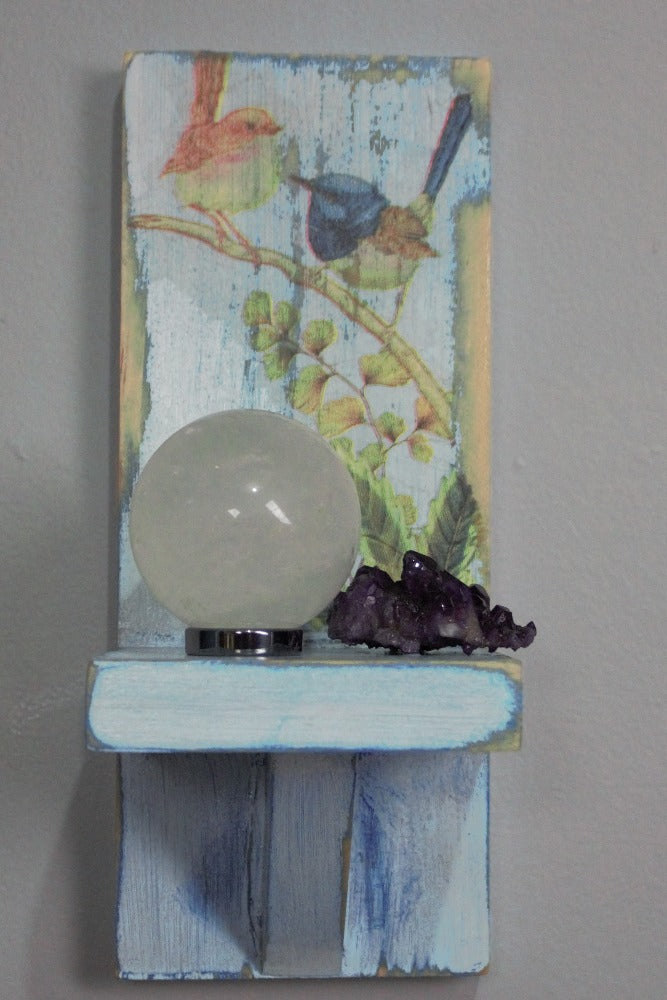 unscented white glass clear quartz crystal ball sitting on a hematite ring next to a purple crystal cluster on a distressed blue and white wooden wall mounted candle or ornament shelf decorated with vintage style brown and blue wrens sitting on a branch with green ferns and blue flowers