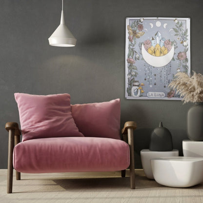 wall hanging of a crescent moon with points facing upwards, crystals and flowers inside the space between the points. The rectangular tapestry is bordered  with vines, flowers and crystals. Tapestry is hanging on a grey wall behind a wooden chair with pink cushions. in front of the tapestry is 3 white abstract tables, 2 contain dark grey vases and 1 has brown grass in it. There is a white light hanging from the ceiling