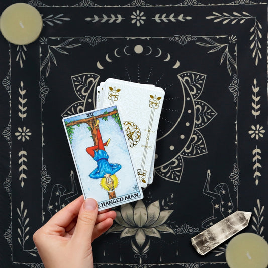 Tarot reader laying out tarot card of the hanged man onto a black and white tarot cloth. a beeswax tealight candle in opposite 2 corners with a clear quartz crystal placed in one corner